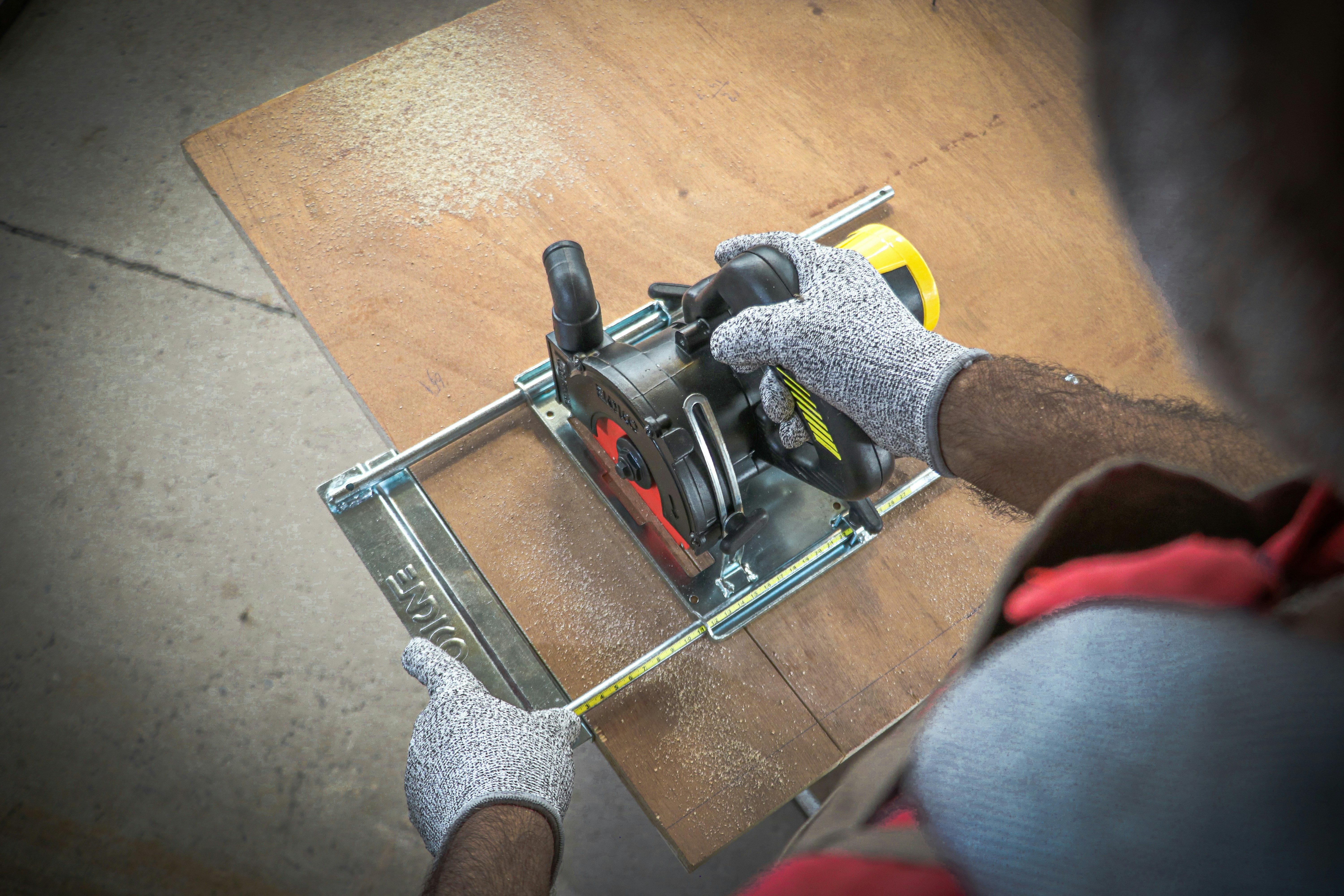 Un ouvriez de l'entreprise du Graveur Signalétique en Martinique; utilise une scie circulaire pour couper du contreplaqué, tout en portant des gants de protection. De la sciure de bois est visible, indiquant un travail du bois actif, concentré et précis.