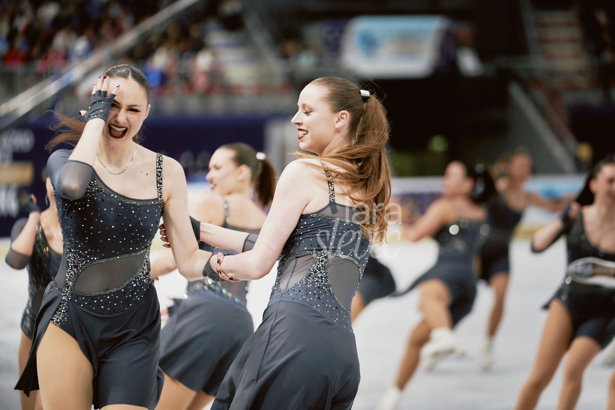 Zwei Synchroneiskunstläuferinnen, die sich am Arm in einer Formation halten. Eins Eiskunstläuferin hält im Lachen ihre Hand vors Gesicht. Team United Angles. ISU Synchronized Skating Championships 2026 in Salzburg. Fotografiert von Lara Slevec.