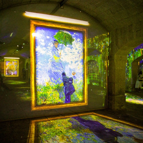 Uma grande pintura emoldurada de uma mulher com um guarda-chuva é projetada em uma parede de pedra em uma galeria de arte, com outra projeção no chão.