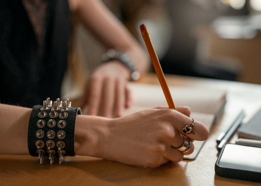 Close up of a person holding pencil, wearing studded bracelet and ring, near a tablet.