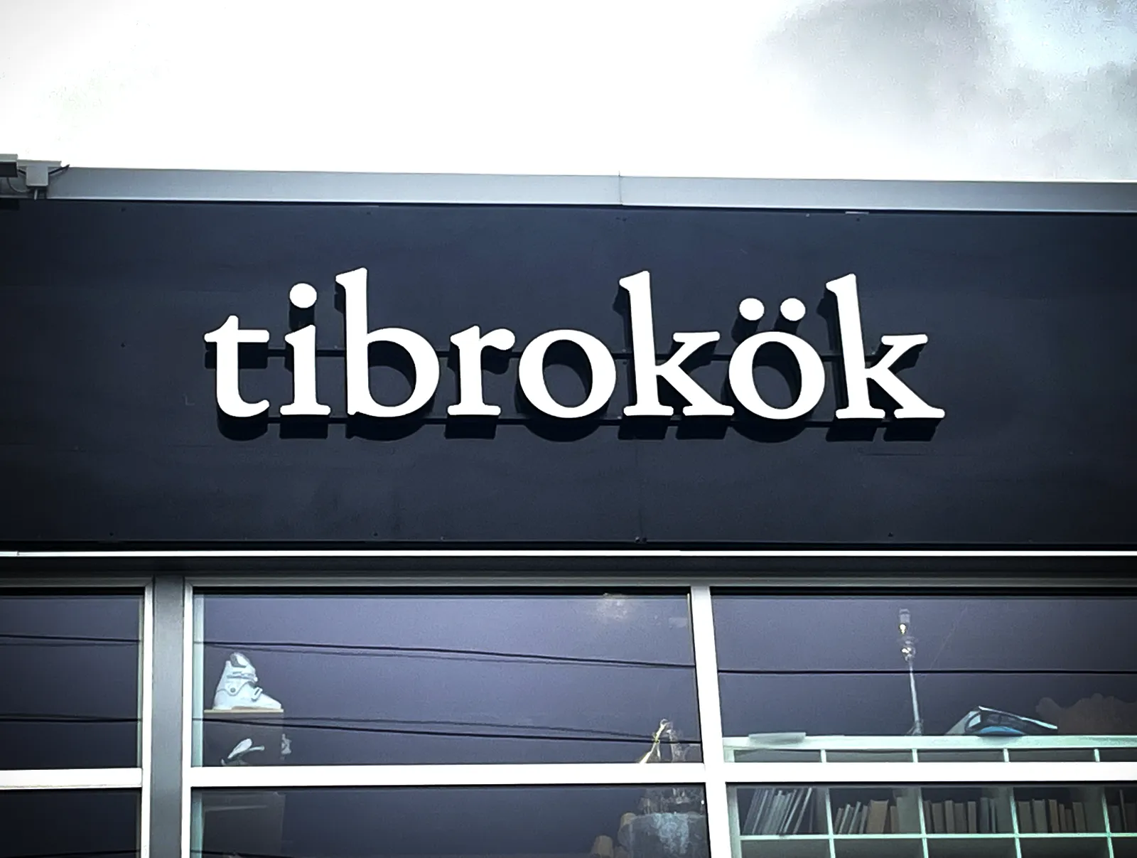 Hvite frittstående bokstaver med teksten tibrokök på svart fasadeskilt over butikkvinduer