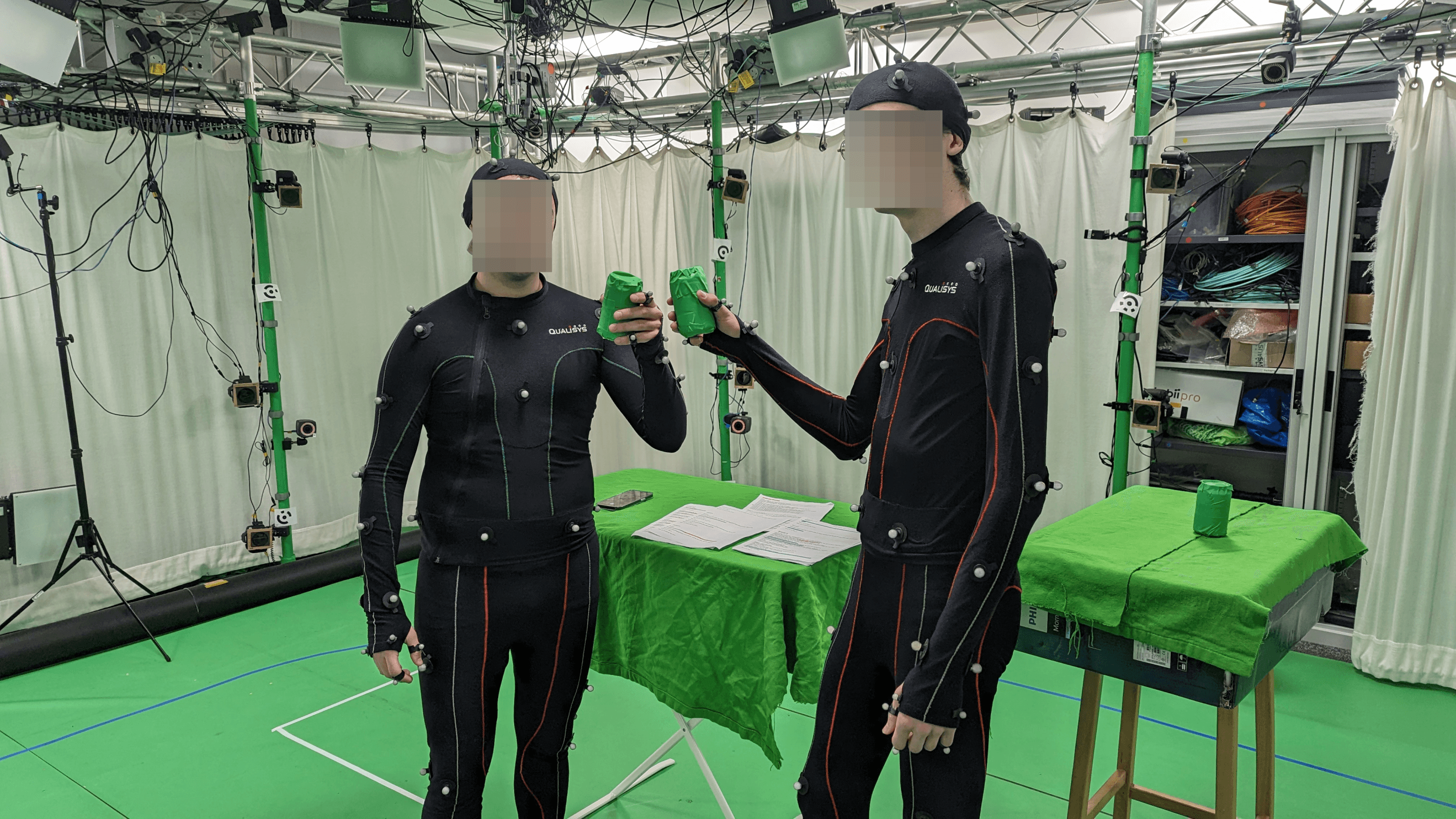 Two actors wearing motion capture suits with reflective markers, toasting with props in a green-screen environment at CAMERA Lab, used for creating realistic VR animations.