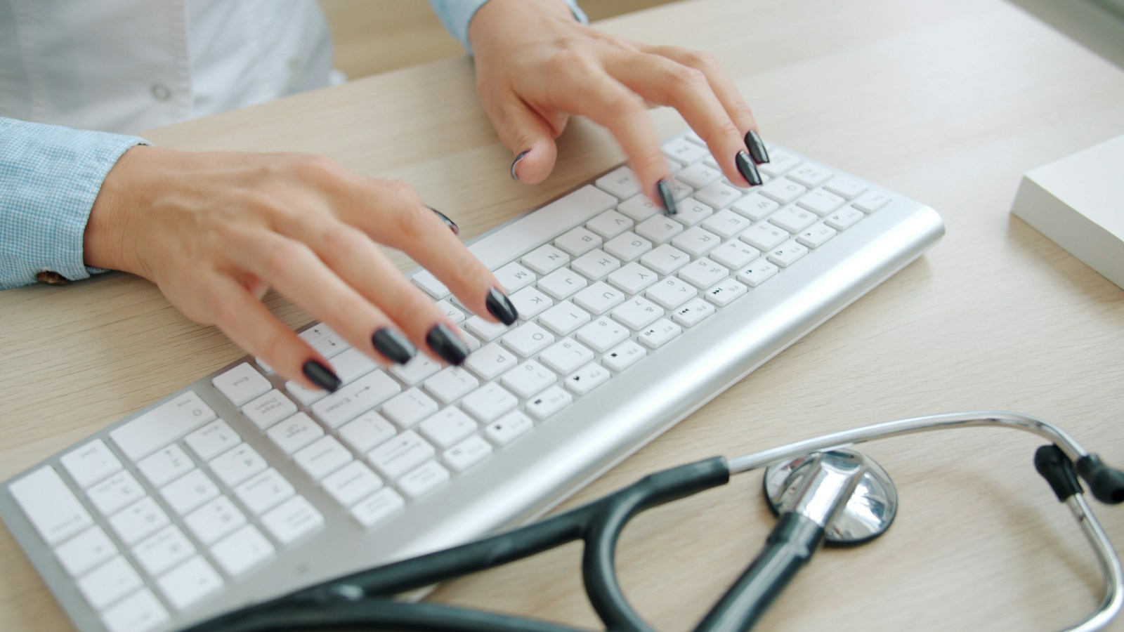 Mains sur un clavier blanc, à côté d’un stéthoscope posé sur un bureau.