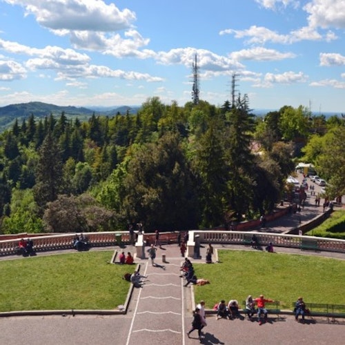 Le persone sono sedute e passeggiano in un'area del parco con spazi verdi, sentieri e una vista panoramica di colline e alberi sotto un cielo parzialmente nuvoloso.