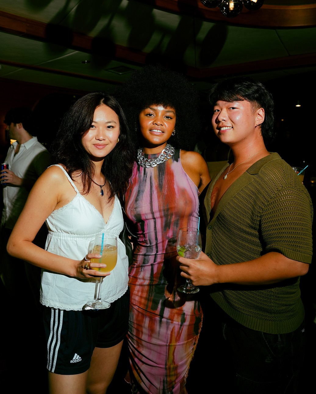 Three people pose for a photo at a social event, smiling and holding drinks, with vibrant lighting in the background.