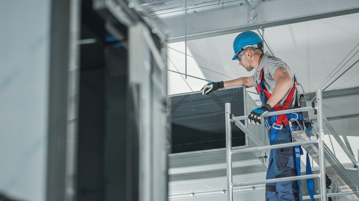 A technician in a hard hat and harness working on ductwork from a mobile scaffold