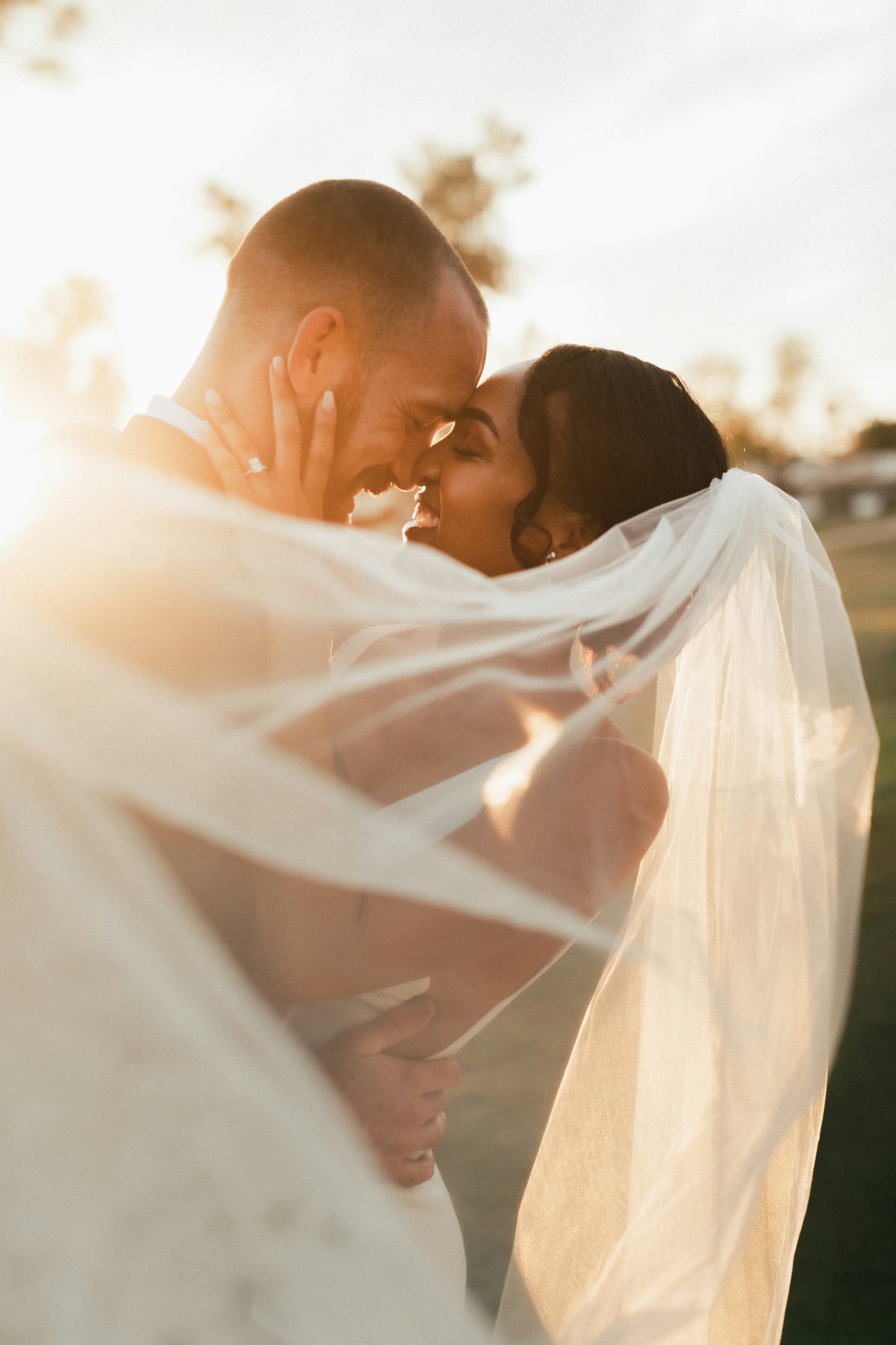 Bride and groom sharing a close embrace wrapped in a flowing veil with warm golden hour sunlight glowing behind them