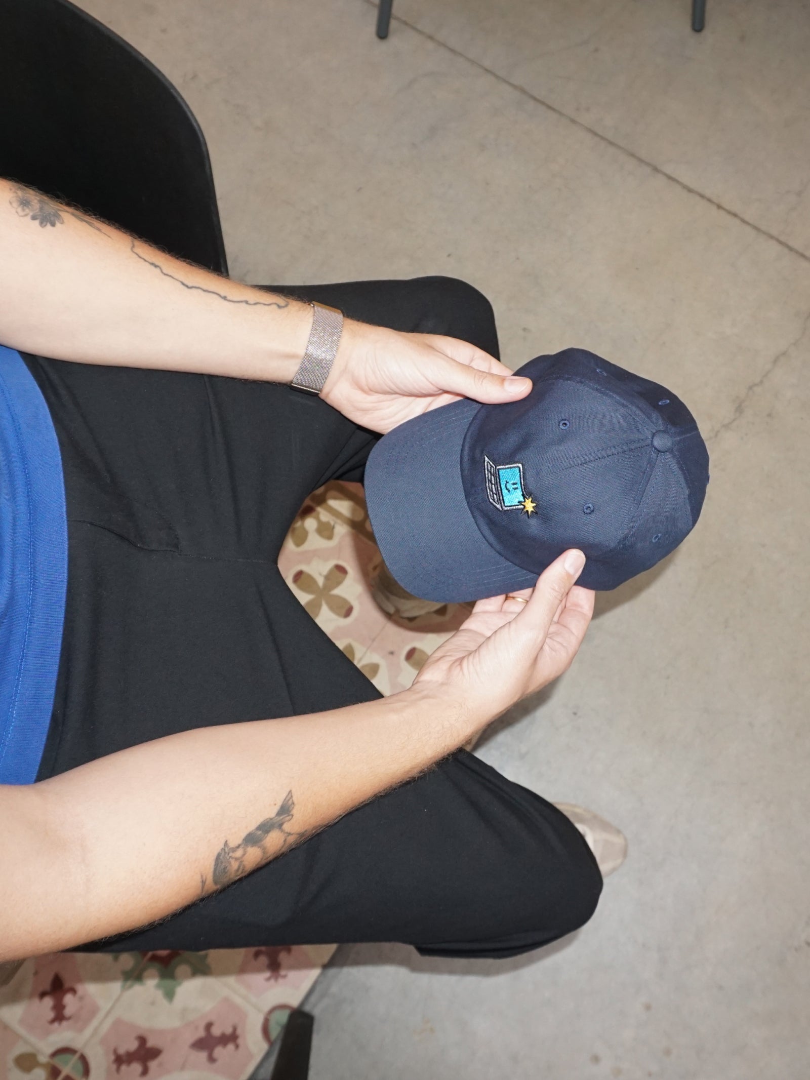Person holding a navy blue cap with a logo on a patterned floor.