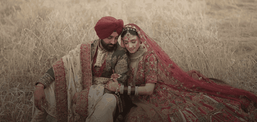 Man and woman sitting in a field, wearing burgundy wedding attire with light brown and gold accents; the man adds a green vest for a vibrant, elegant touch.