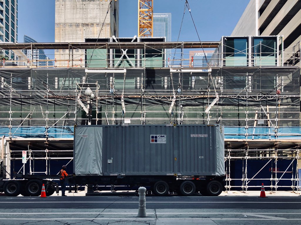 A large flatbed truck stationed at the construction site, carrying a guestroom module ready for installation atop the already stacked units.