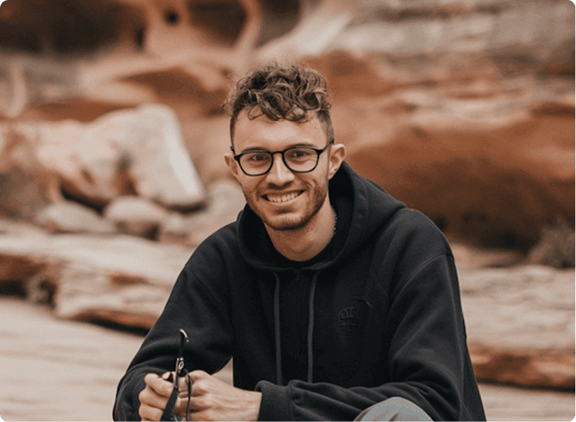 A person with glasses and curly hair sits smiling in front of a blurred, rocky desert landscape, holding a pair of sunglasses, conveying a sense of adventure and outdoor exploration.