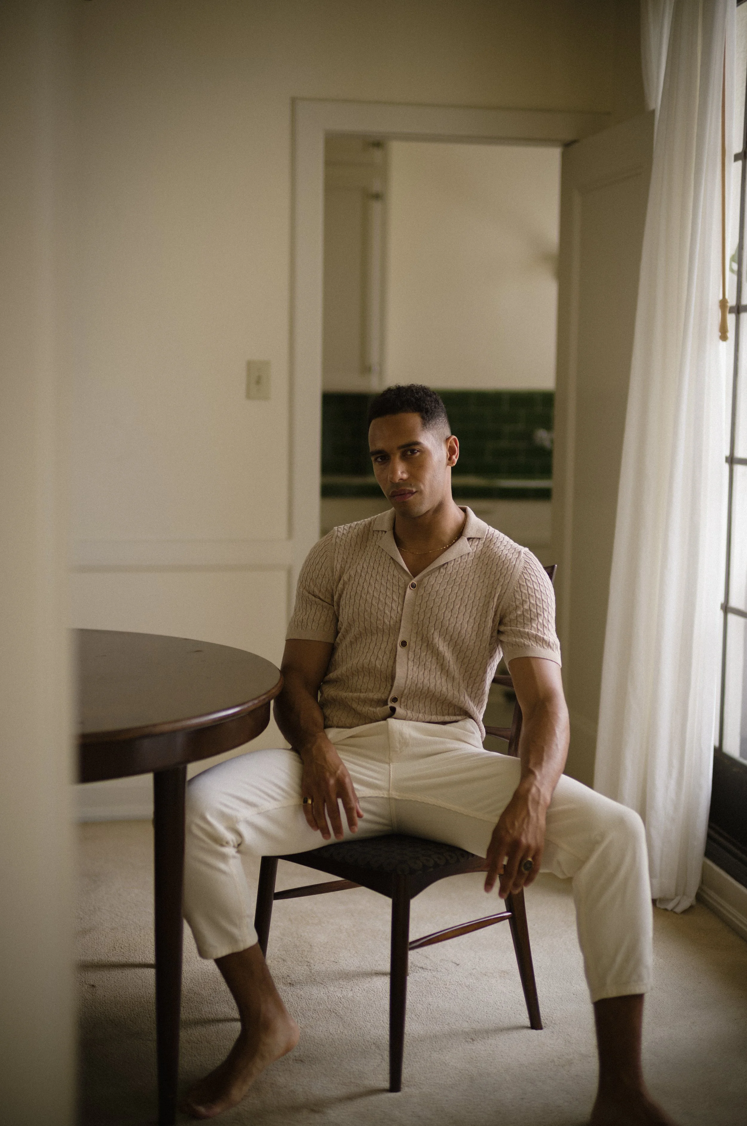 Man in a cream cable-knit shirt and trousers sitting astride a wooden chair in a sunlit, sparsely furnished room.