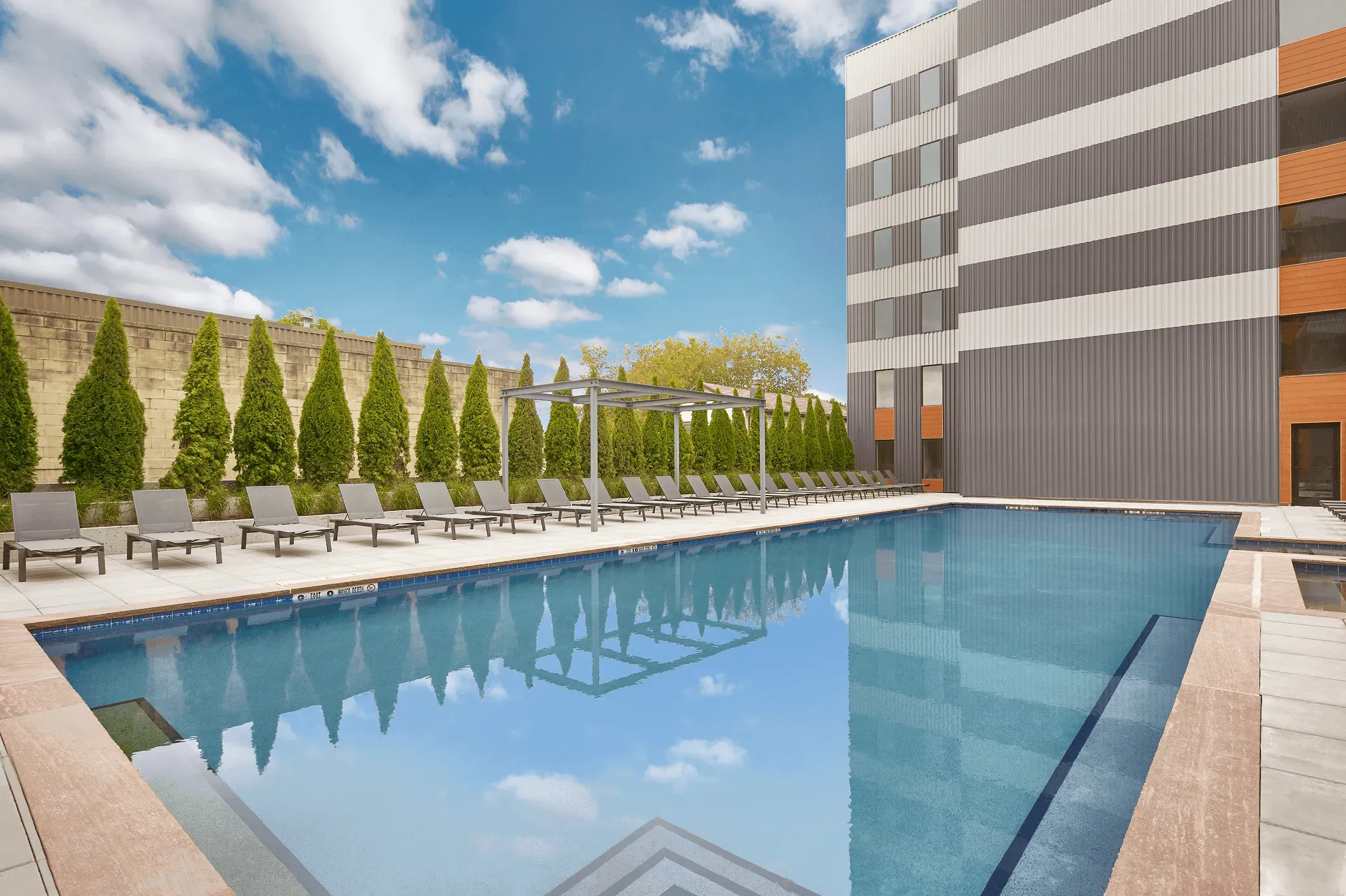A clear swimming pool surrounded by lounge chairs and greenery, with a modern building in the background under a blue sky.
