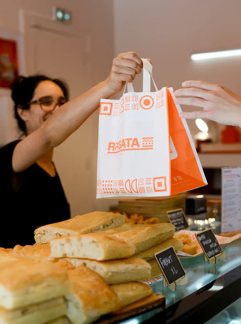 Sac à emporter Rosata personnalisé aux couleurs de la marque, remis à une cliente devant le comptoir de focaccias.
