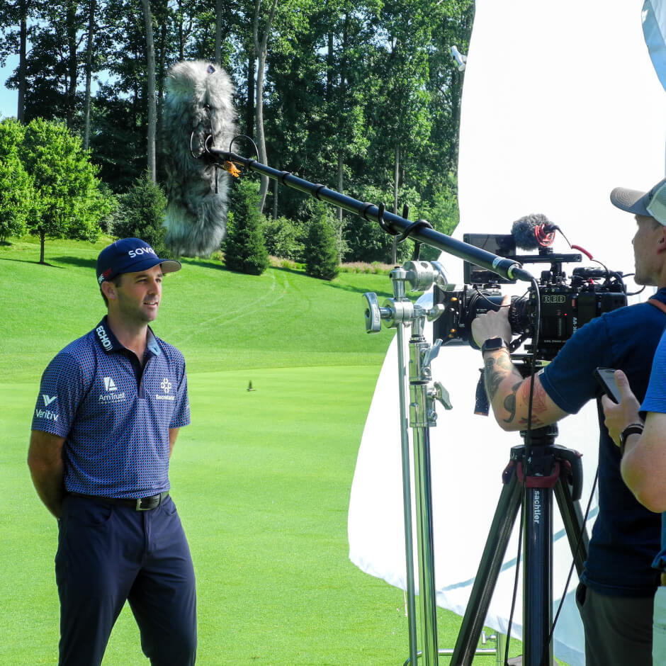 A golfer stands on a golf course being filmed by a cameraman with a boom microphone and lighting equipment visible.