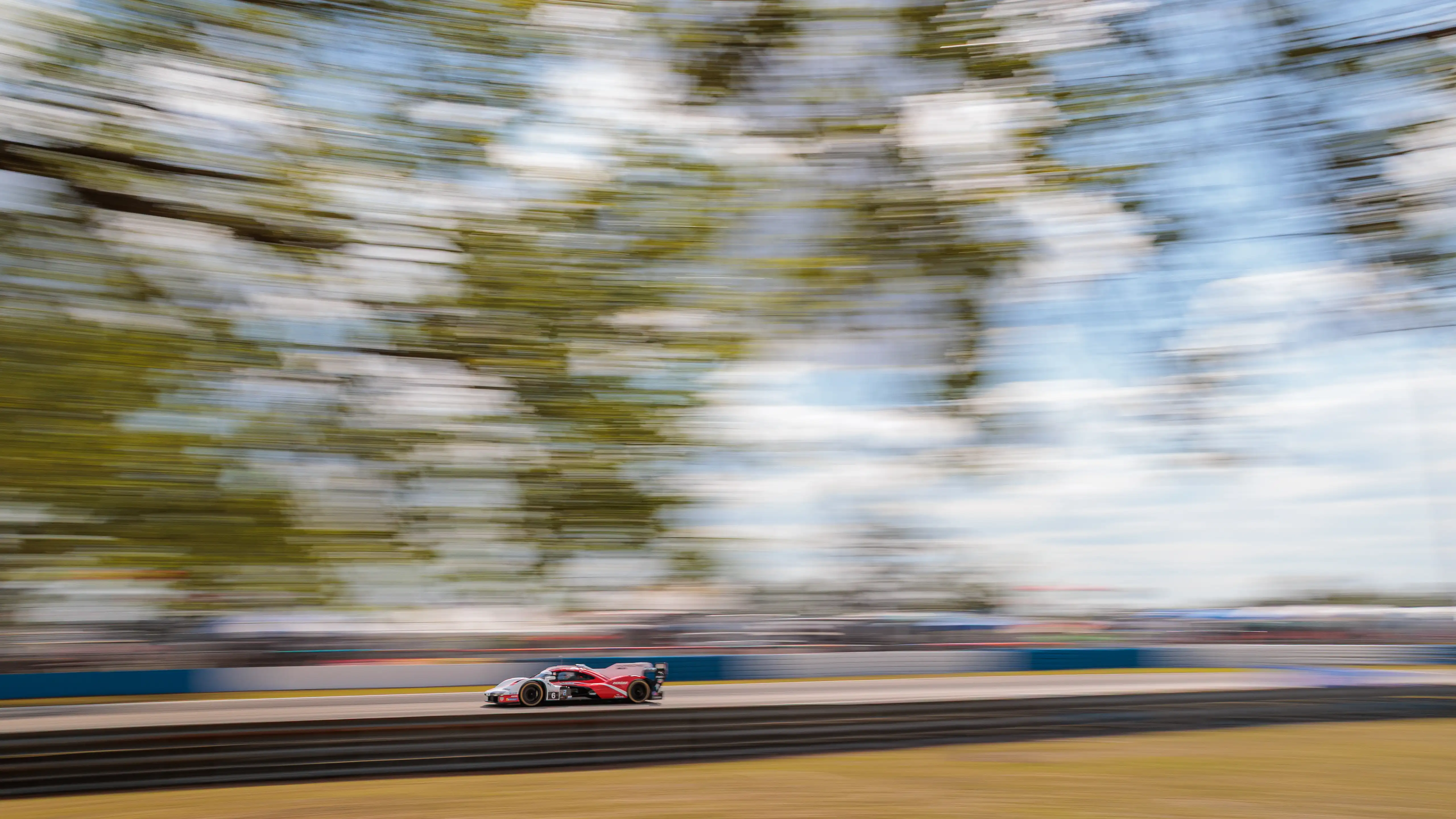 A red Porsche racing car speeds down a track, against a blue sky and trees, with a motion blurred background.