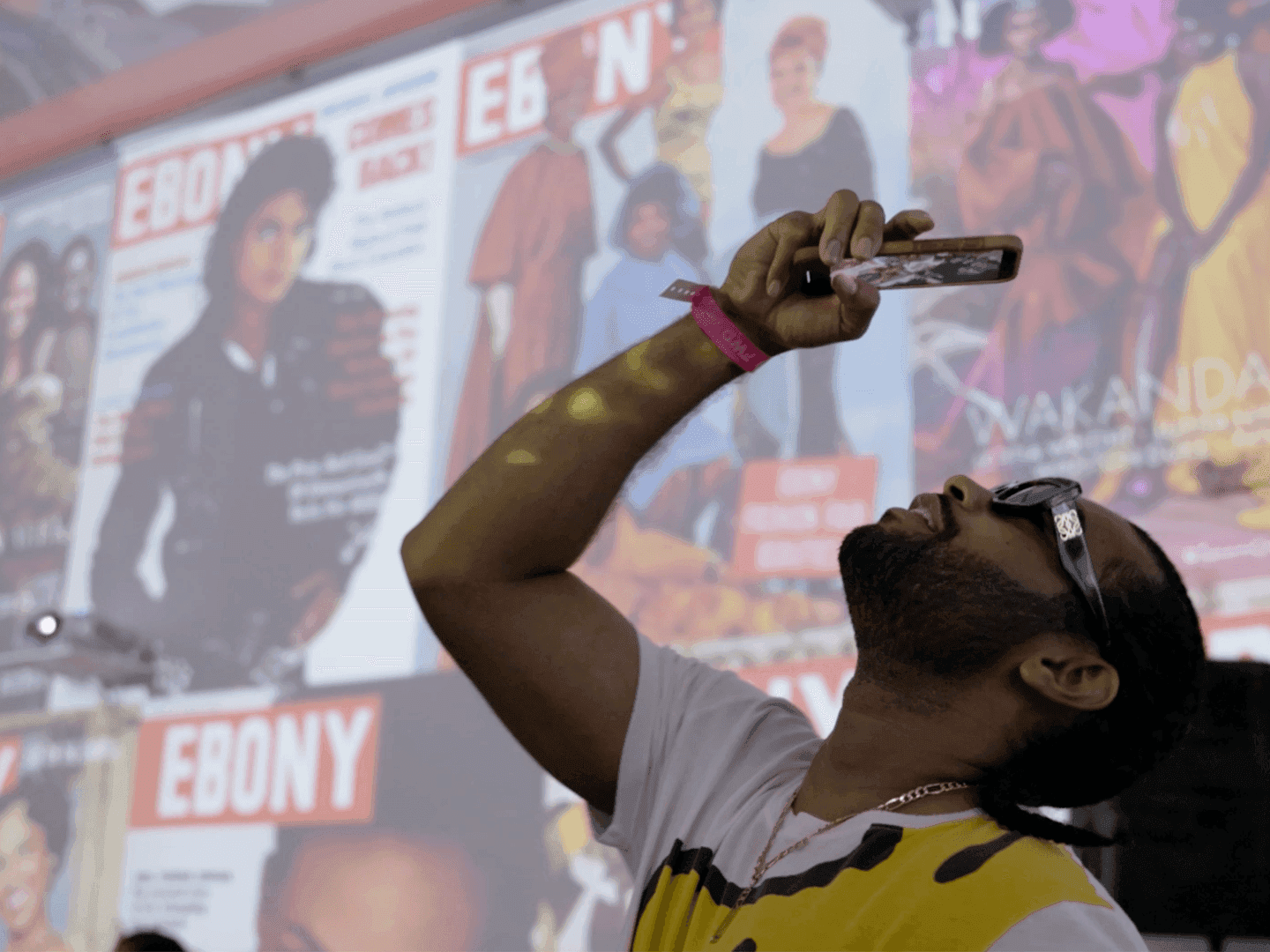 A man wearing glasses and a pink wristband holds up his phone to take a photo, with a large display of vintage Ebony magazine covers projected on a wall behind him.
