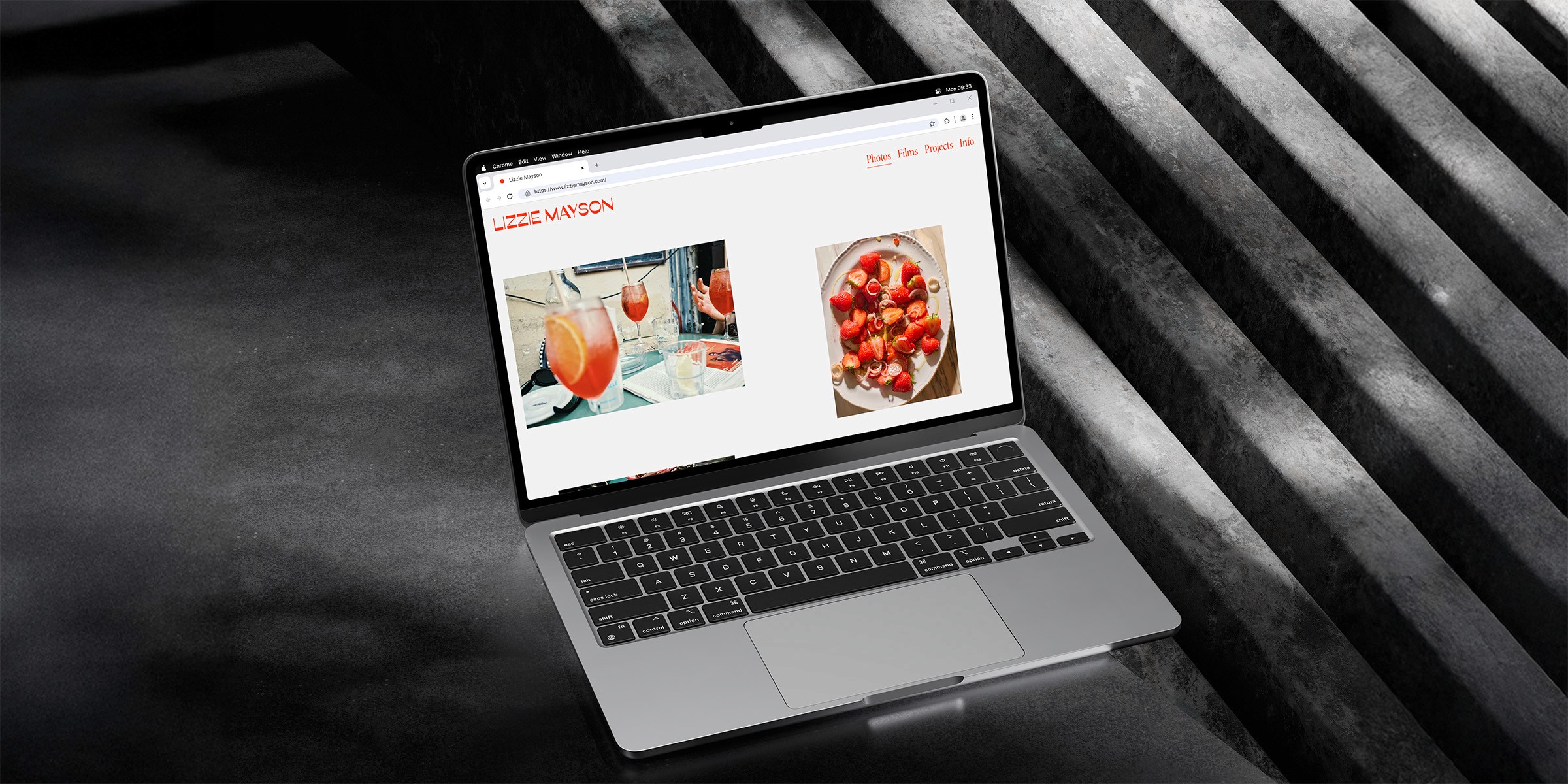 A sleek laptop displays a minimalist photography portfolio of Lizzie Mayson's website, featuring vibrant images of cocktails and a close-up of strawberries, set against a modern, dark industrial background, highlighting a stylish creative workspace.