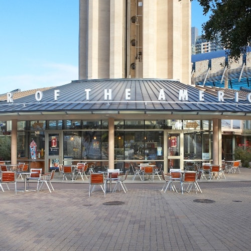Outdoor café with empty tables and chairs in front of a glass-walled building labeled "OF THE AMERIC." Towering structure behind.