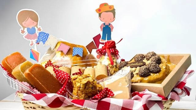 Cesta de vime com paçocas, bolo e outros doces típicos, decorada com itens característicos de São João.