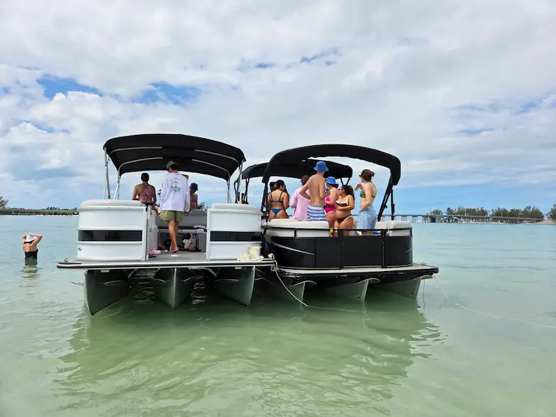 A group of people enjoy a leisurely day on a pair of pontoon boats floating in the clear, calm waters surrounded by scenic views, under partly cloudy skies in Bradenton, FL.
