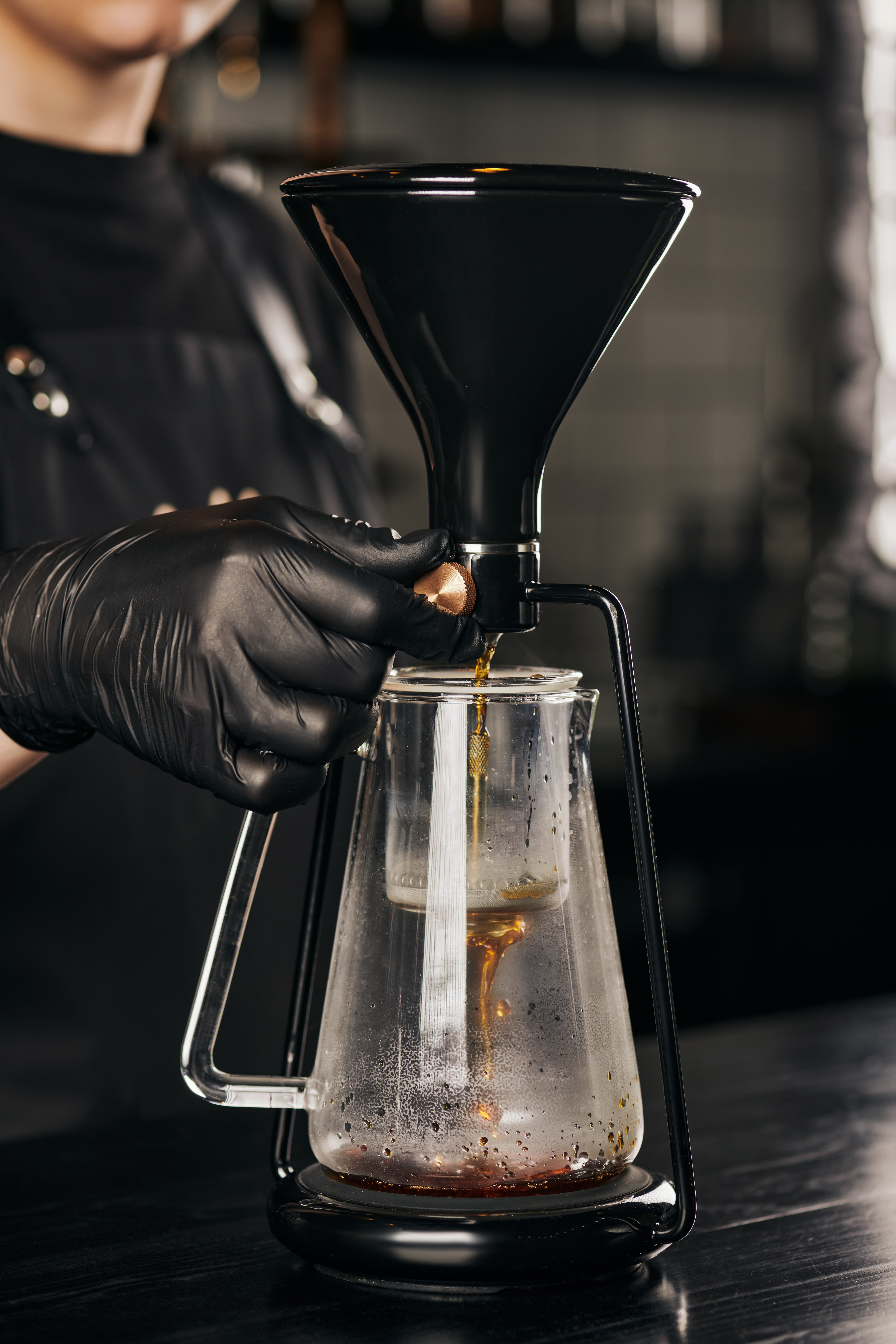 Barista using a siphon coffee brewer as coffee drips into a glass carafe, captured in a dark, moody setting.