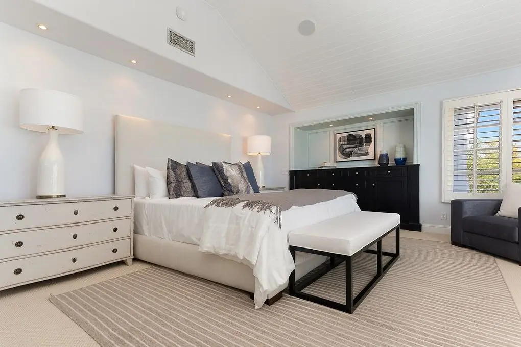 'Port Streets Remodel' serene bedroom with a plush bed, striped ottoman, dark wood dresser, and soft lighting from a large window.