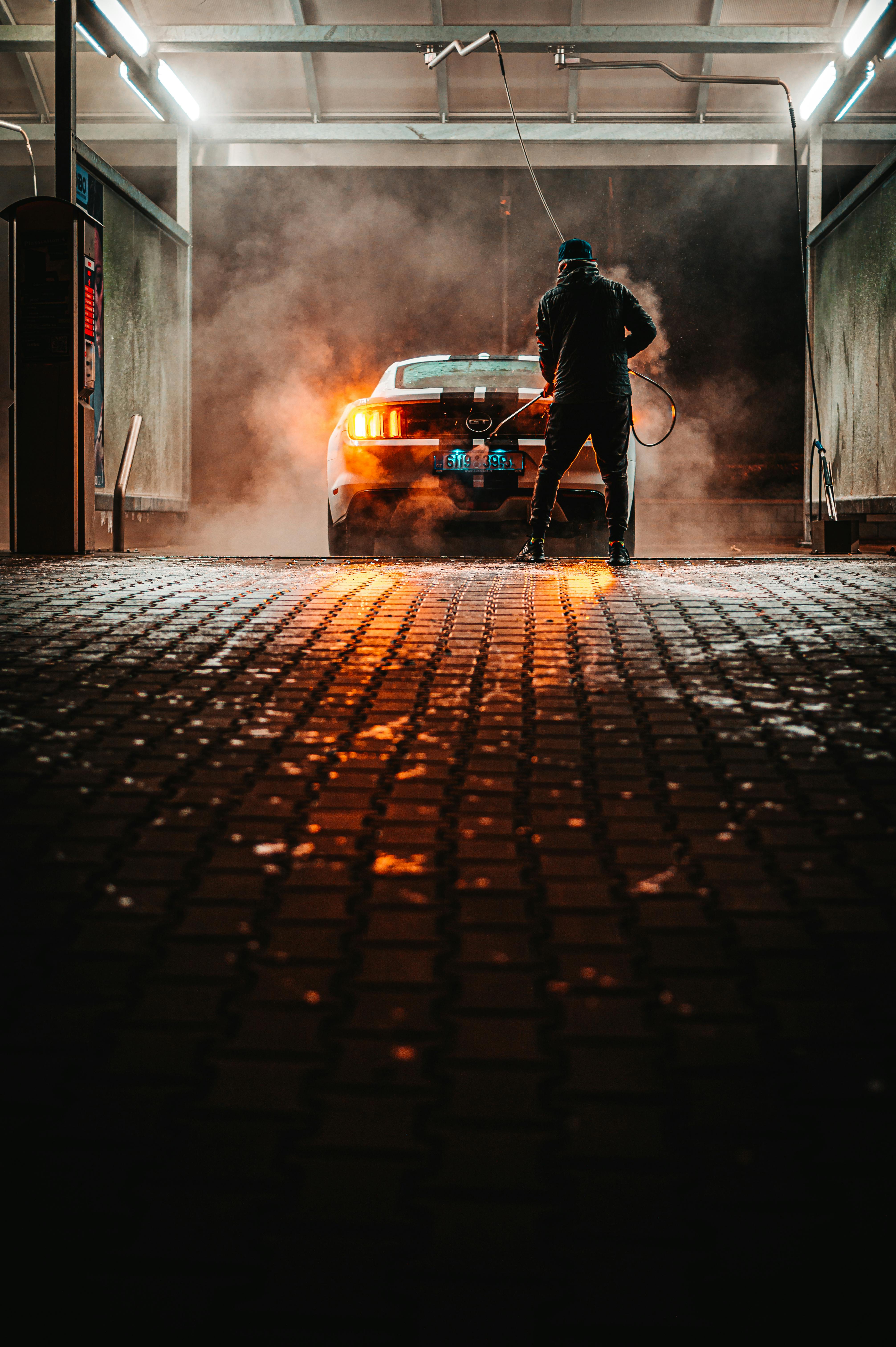 Two people in a workshop, one standing and smiling while the other leans over a car, working on it.