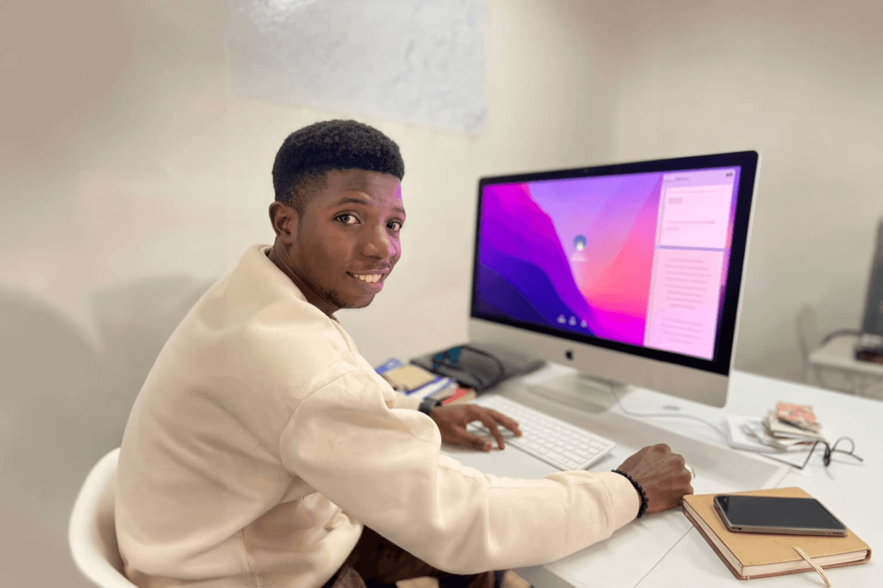 A young man smiling at the camera while sitting at a white desk and using a desktop computer in a bright indoor workspace, with a notebook and phone beside him.