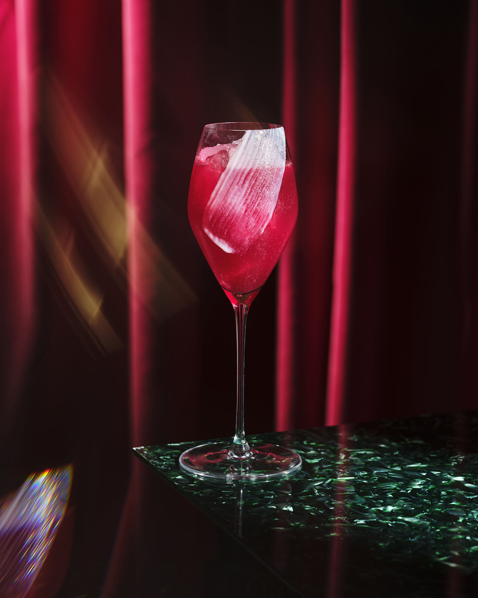 A close-up of a red drink in a glass, illuminated by soft lighting, with colorful beads scattered nearby.