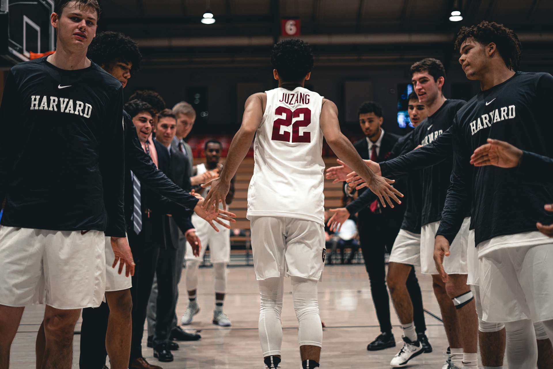 A basketball player in a white jersey runs onto the court, high-fiving teammates wearing black long-sleeved "Harvard" shirts.