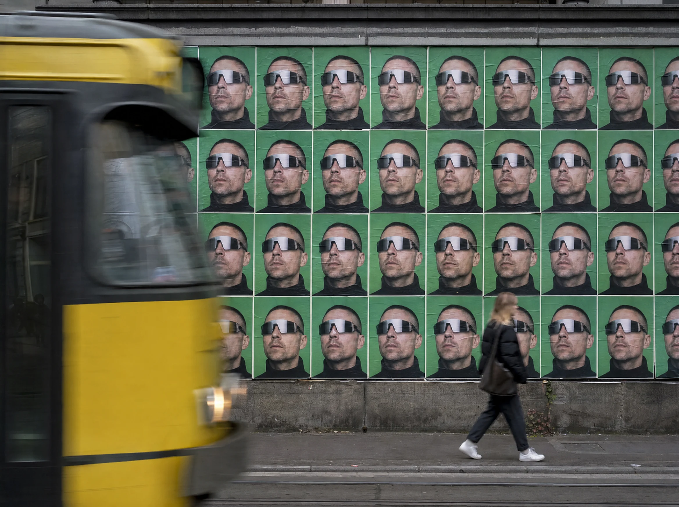 Wall of pasted posters of a man in chrome sunglasses on green, with a tram passing