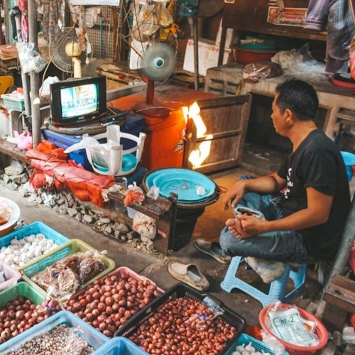 남성이 양파, 마늘 및 기타 농산물 바구니로 둘러싸인 시장 가판대에서 작은 TV를 보고 있습니다.