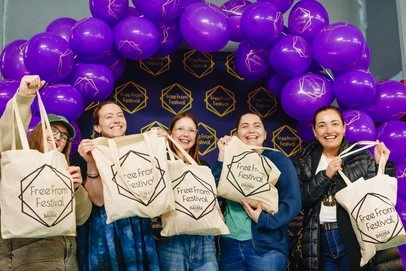 group of women holding up their free from festival goodie bag up and smiling infront of balloon arch