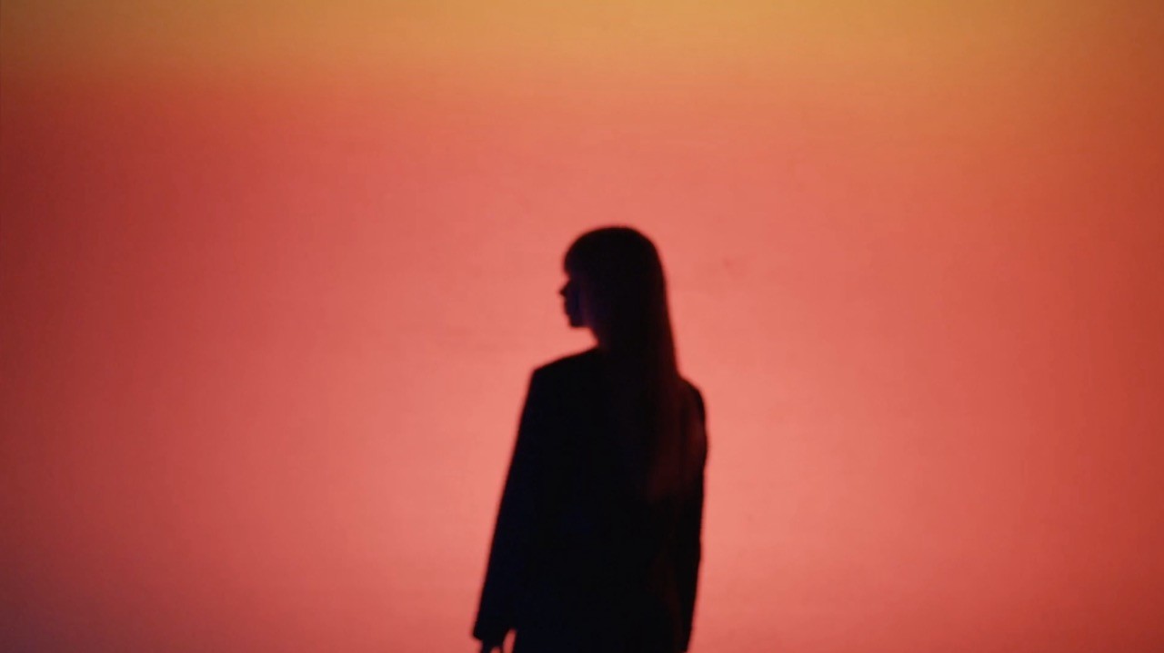 Silhouette of a woman with long hair standing against a warm orange-red gradient background.