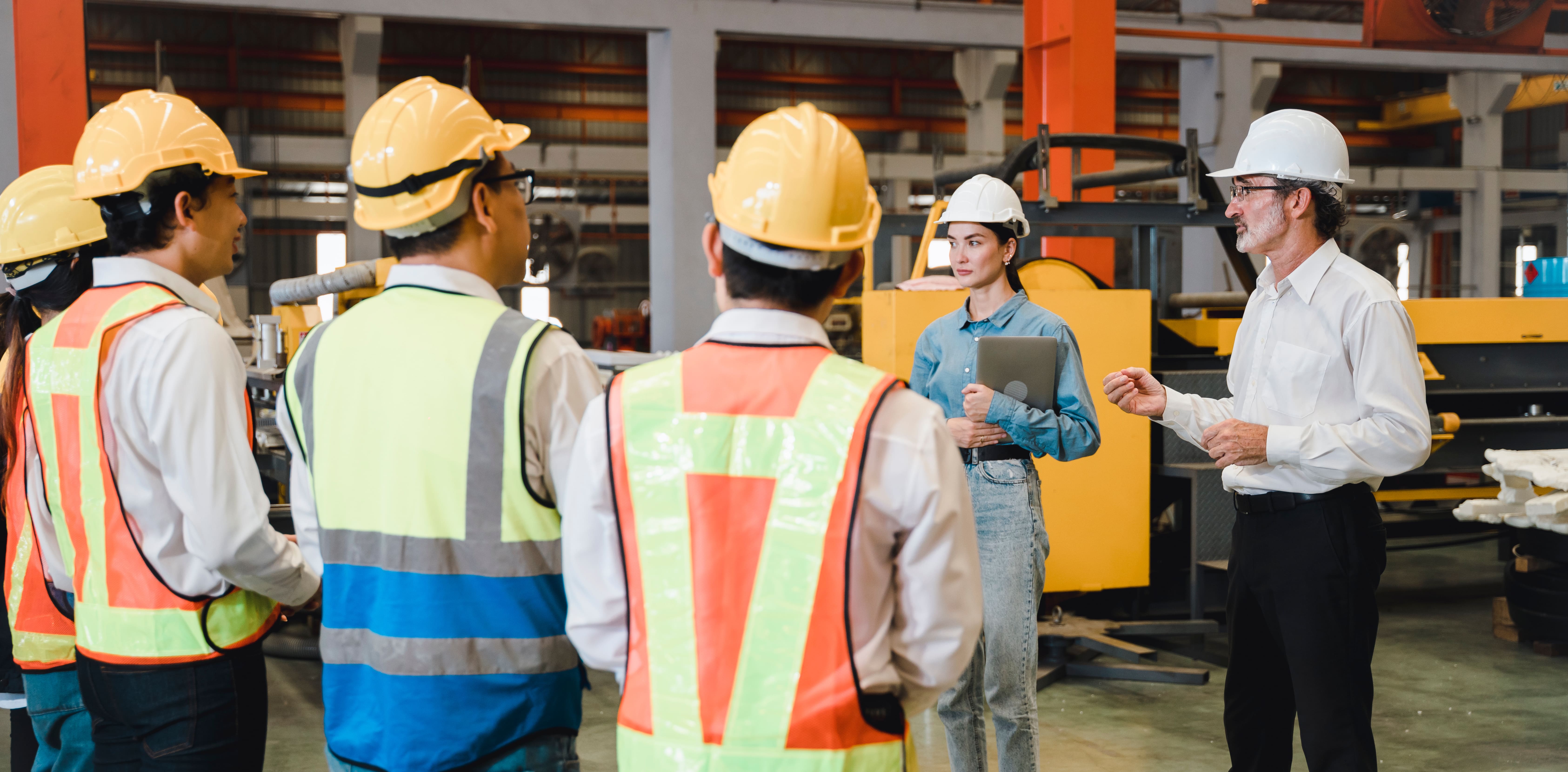 A team of workers wearing hard hats and safety vests listening to a supervisor in an industrial facility.