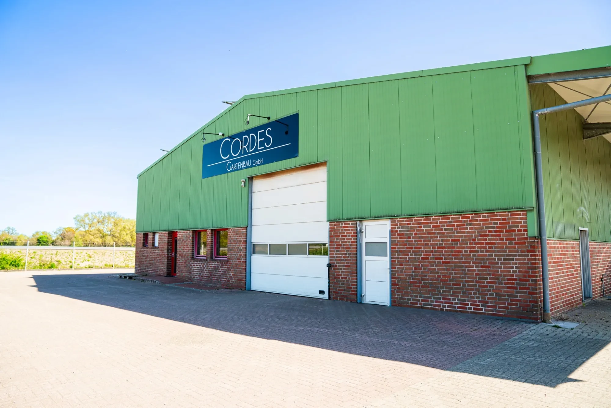 Grüne Lagerhalle der Cordes Gartenbau GmbH mit Firmenschild und gepflasterter Hoffläche