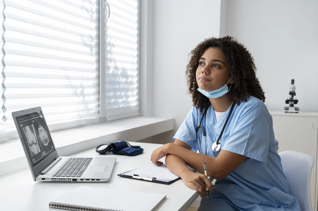Young resident doctor wearing light blue scrubs, sitting at a desk, looking off in the distance.