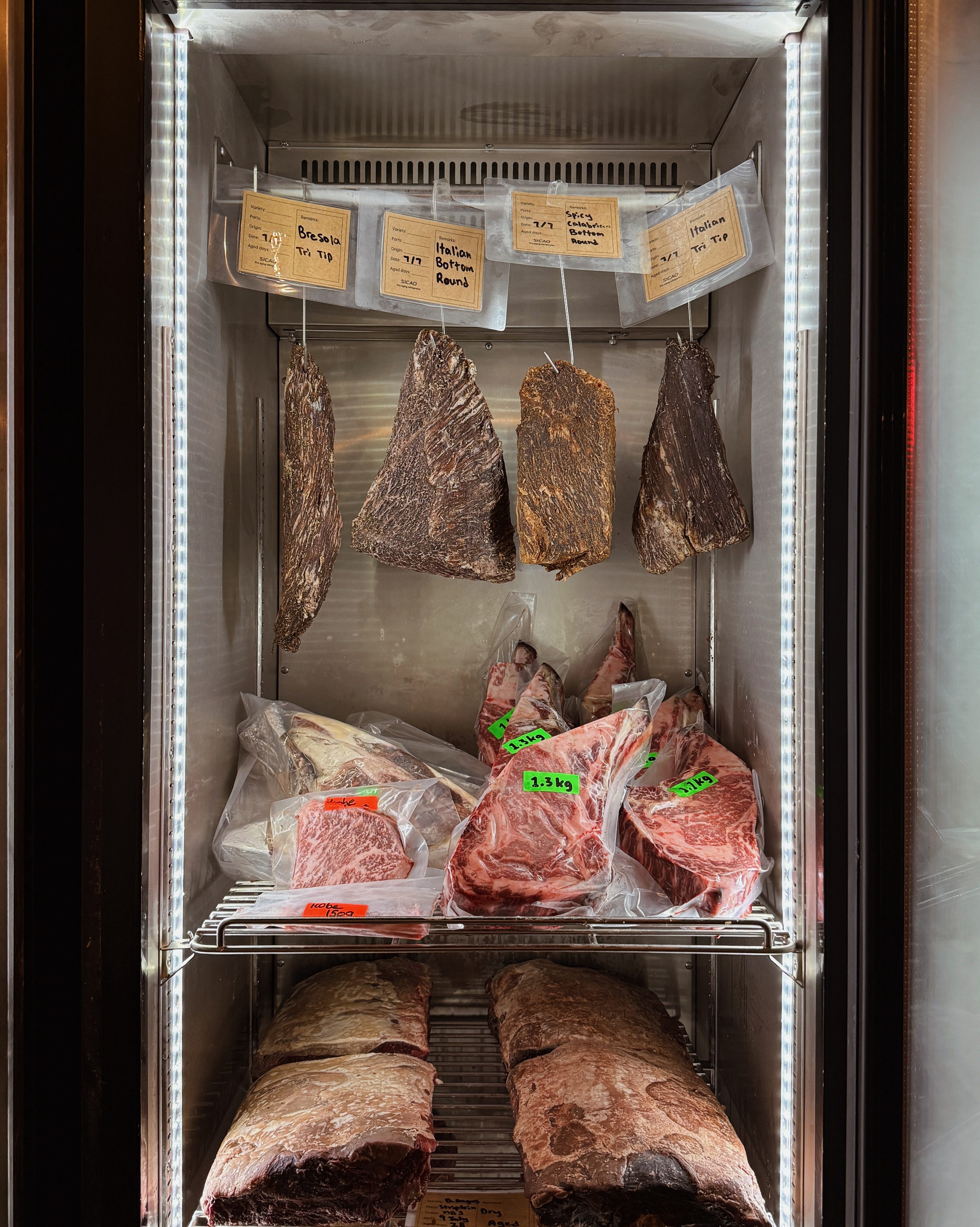 An organized display of various cuts of meat stored in a refrigeration unit, showcasing dry-aged beef with labels indicating their names and weights.