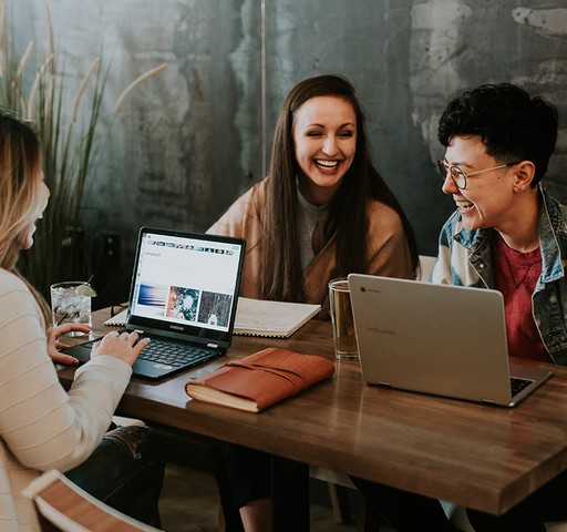 Three individuals engaged in a friendly meeting at a wooden table with laptops, notebooks, and drinks, in a room with ambient lighting and greenery in the background