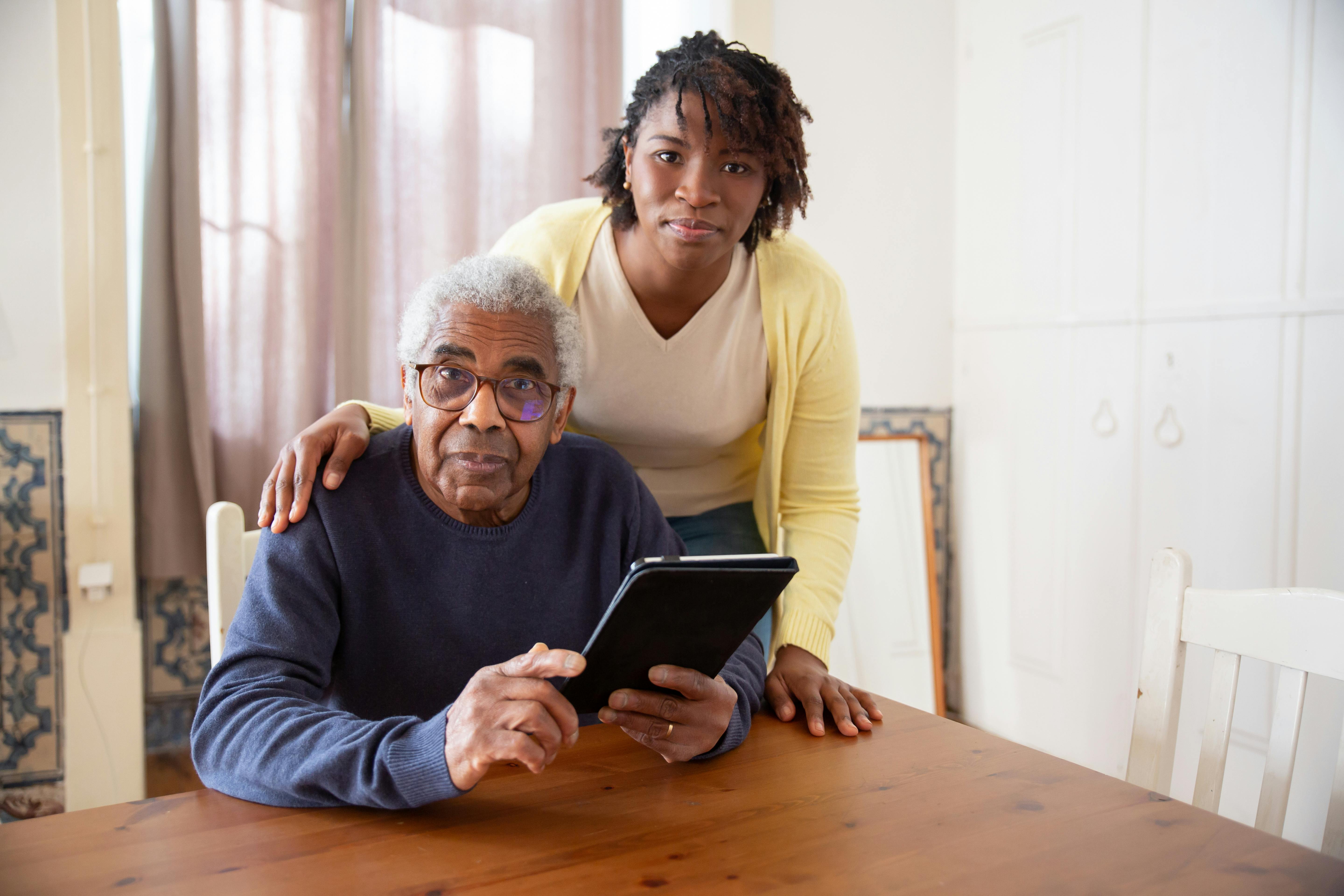 An older black man sits at a table holding an ipad with a younger woman standing behind him with her hand on his shoulder.