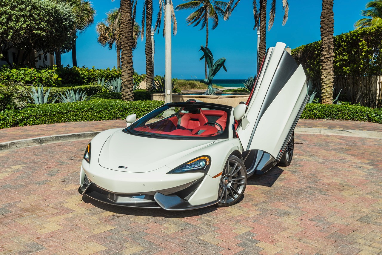 Front view of a white McLaren 570S with its dihedral doors open, parked in a tropical setting with palm trees in the background.