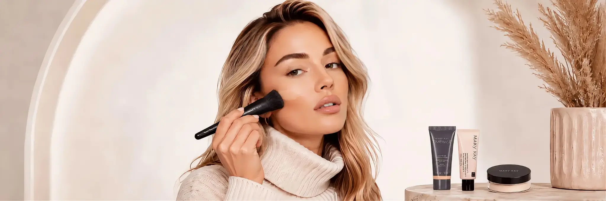 Modelo loira usando blusa de gola alta clara e aplicando maquiagem com pincel, com produtos Mary Kay ao lado.