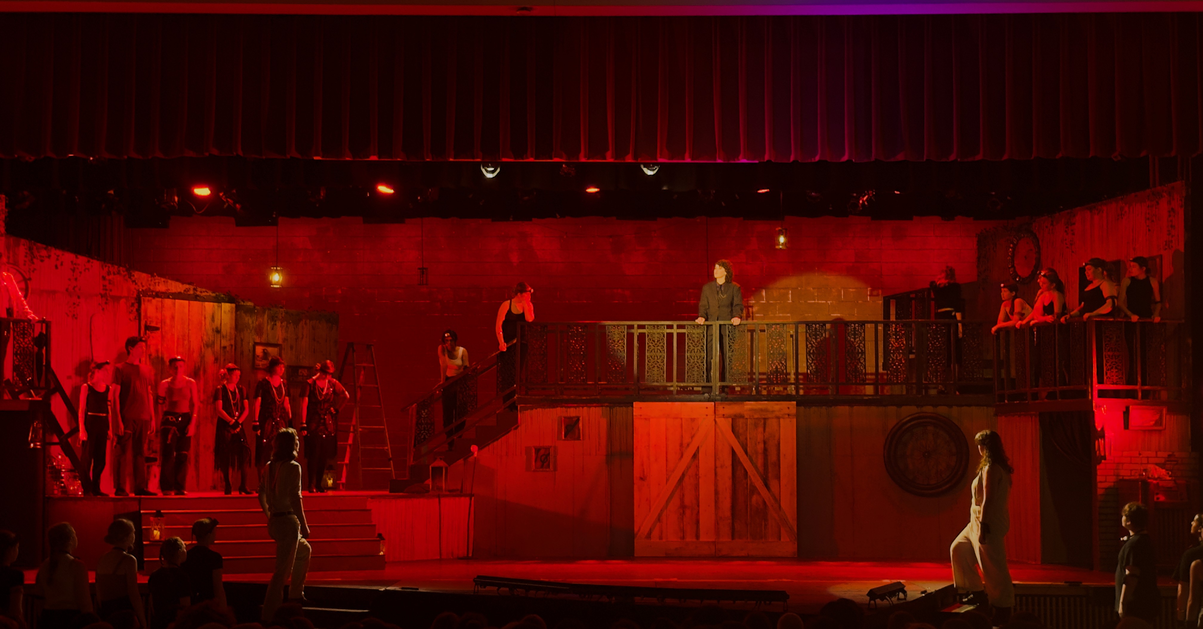 The stage is set with saturated red lighting, creating a dramatic ambiance as Hades stands prominently on an elevated platform in a spotlight. Orpheus and Eurydice, with workers behind them, look up at him. They are lit with backspots that reveal their form while keeping Hades the focus of the scene.