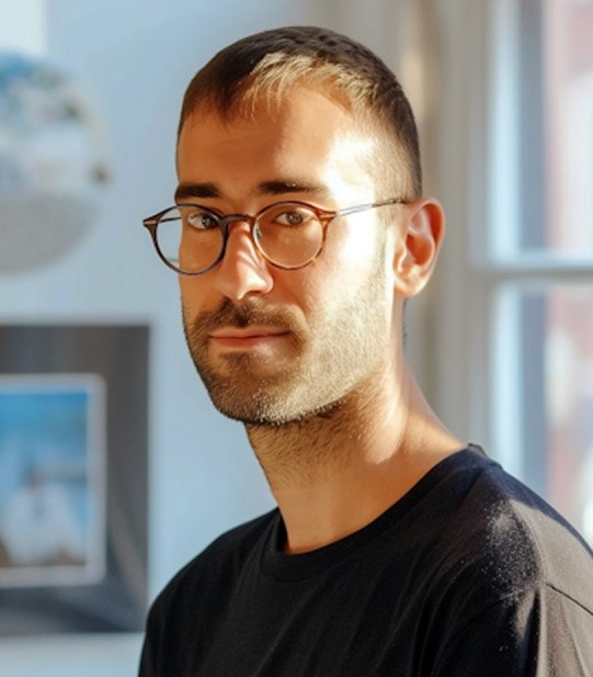 In a warmly lit room, a man with glasses stands beside a wall lined with books and framed art, gazing at the camera, creating a serene yet focused atmosphere.