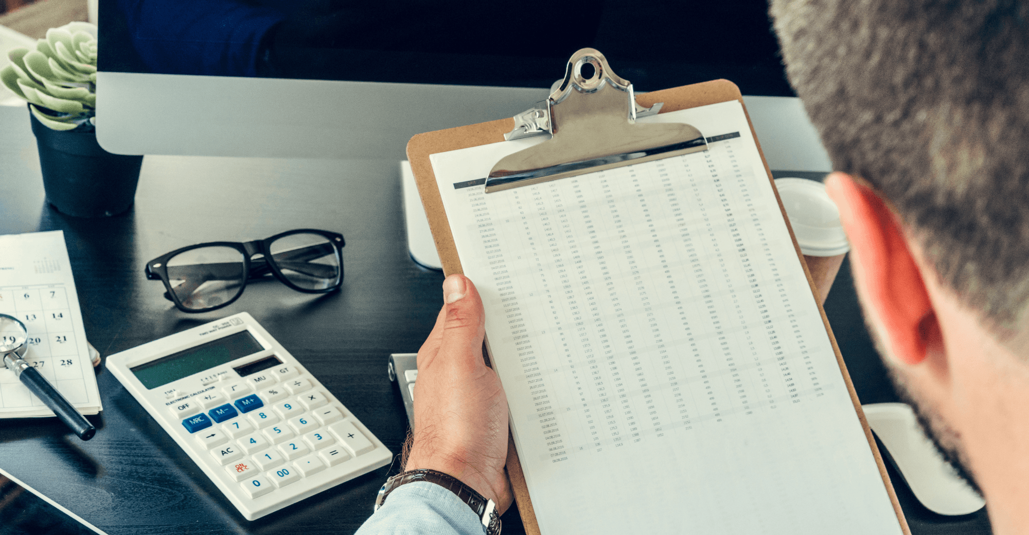 Man holding a clipboard over a desk with a calculator and glasses on it.
