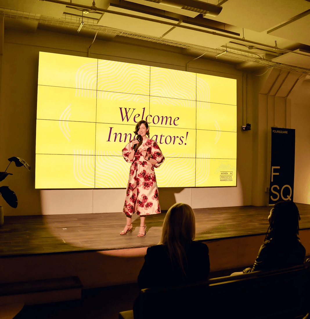 A speaker in a pink dress stands on stage in front of a large screen displaying "Welcome, Everyone!"