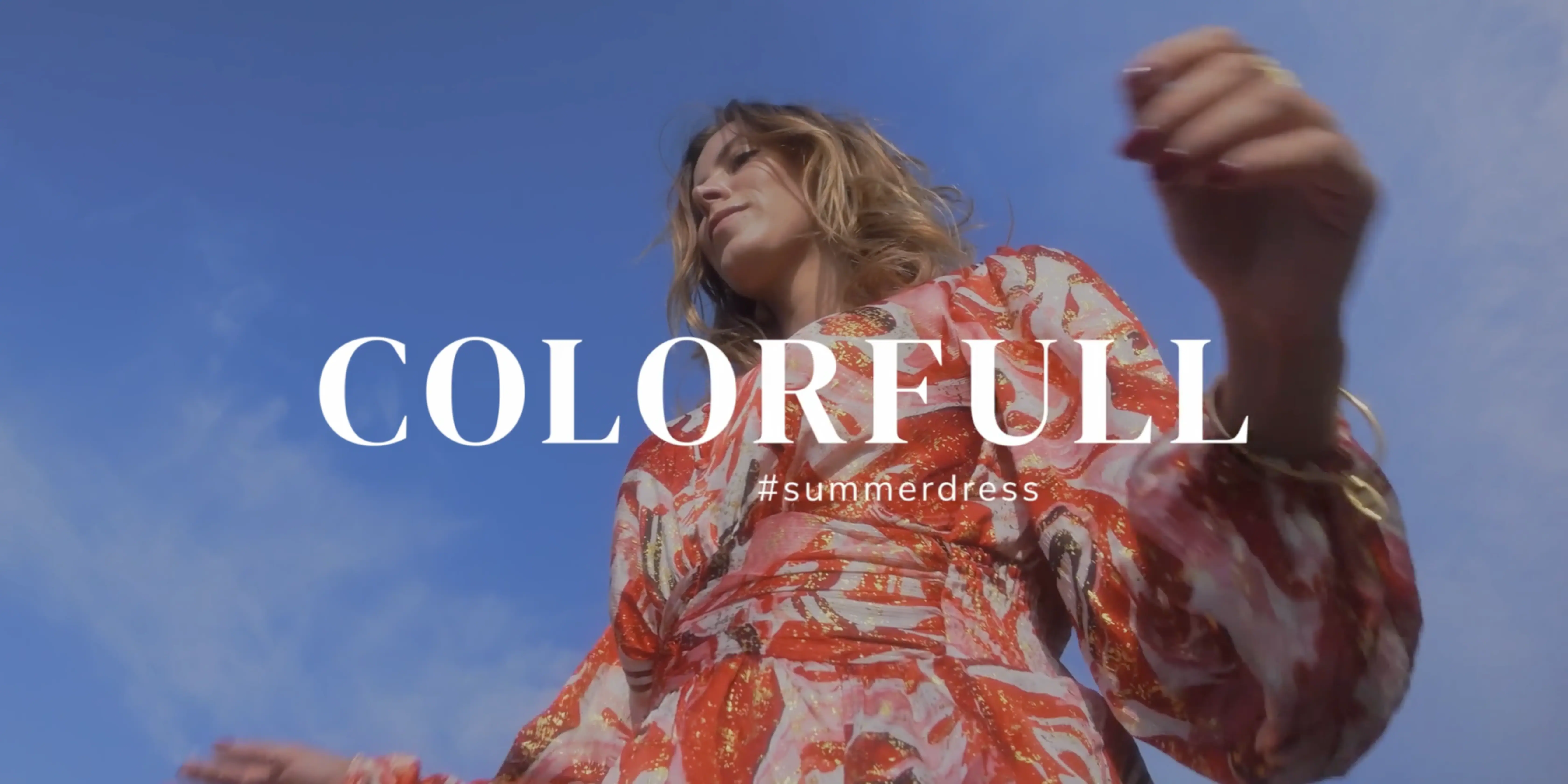 A person wearing a vibrant, floral-patterned summer dress poses under a clear blue sky, embodying a stylish and colorful fashion choice.