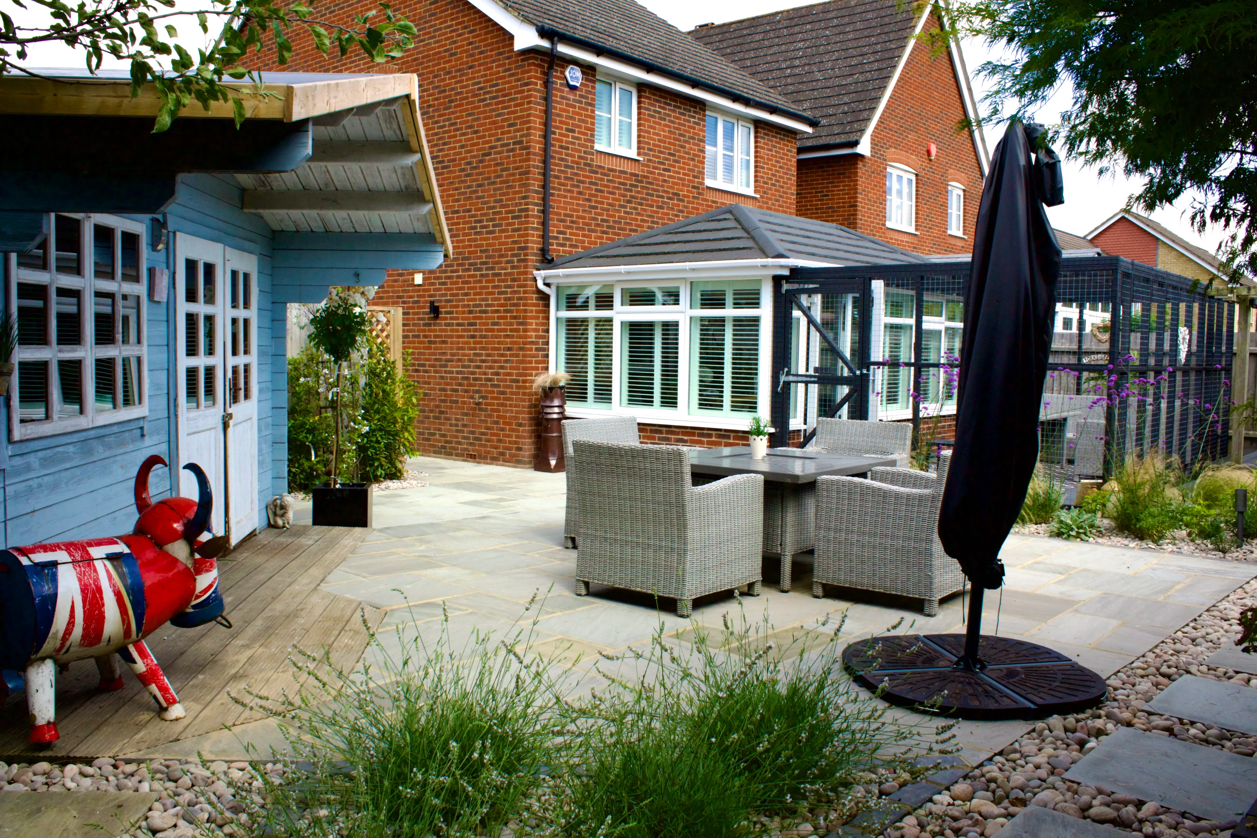 A cozy outdoor patio with seating, a large umbrella, and a blue shed next to a house surrounded by greenery.