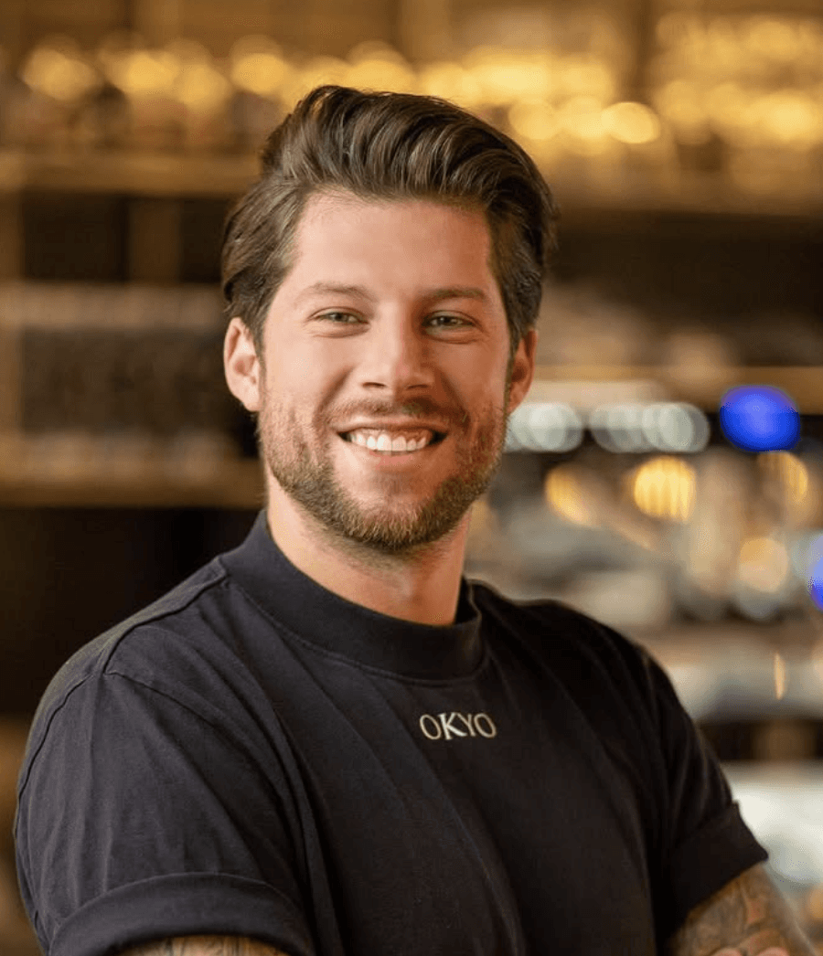 Ein junger Mann mit schwarzem t-Shirt und verschränkten Armen schaut in die Kamera.